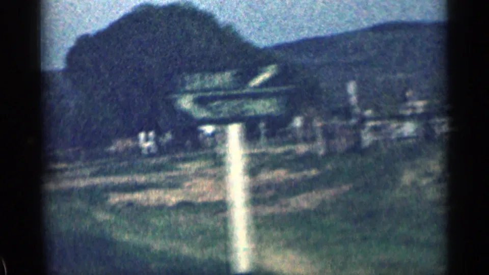 1963 Colorado 8mm view of a roadside signpost, low buildings, open ground, trees, and hills from a moving car.