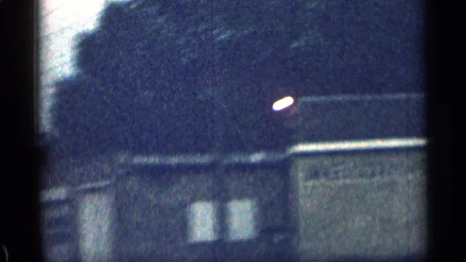 1963 Colorado home-movie frame with sloped roofs, utility wires, and a wooded hillside in soft focus.