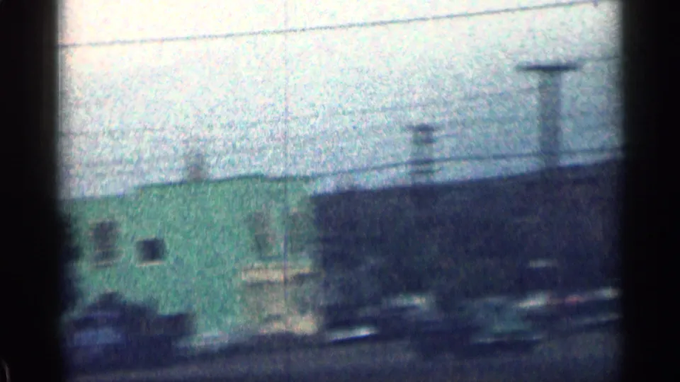 1963 Colorado street view with a green building, utility poles, wires, and blurred vehicles in a grainy 8mm frame.