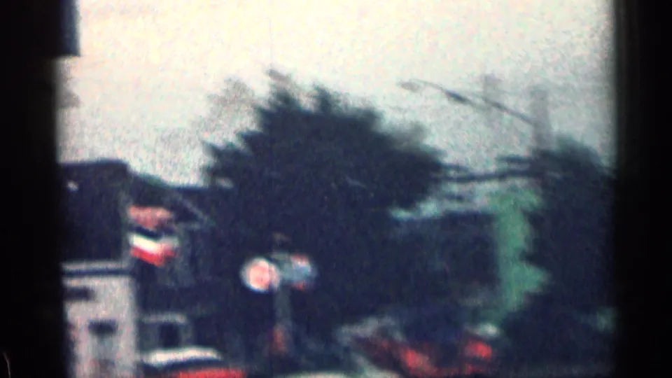 Soft 1963 Colorado 8mm frame showing a blurred suburban street view with foliage, a person in yellow, and a Chevron sign behind them.