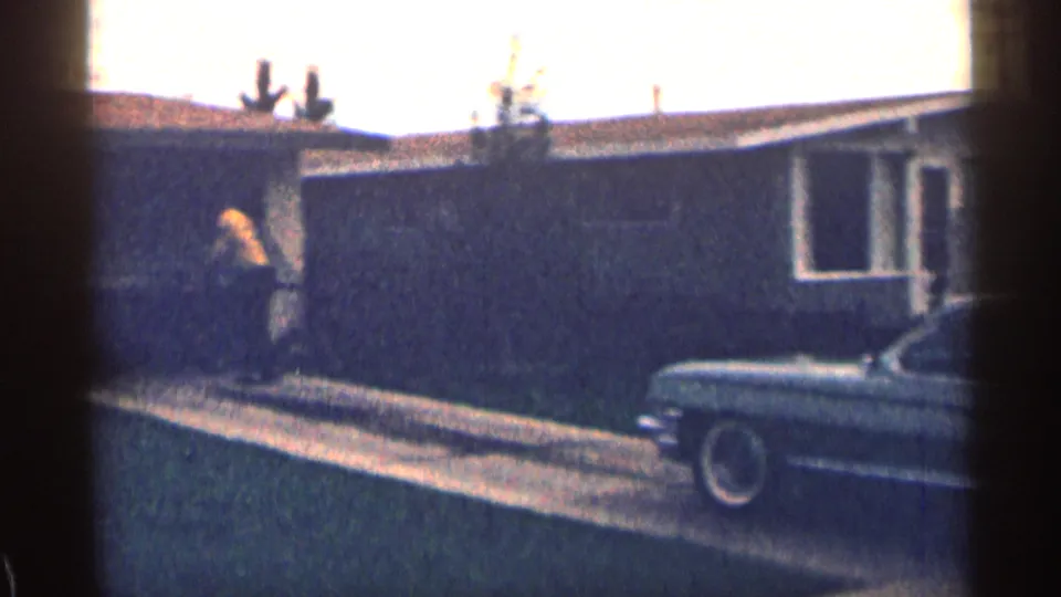 1963 Colorado home-movie frame with a person in yellow near the house wall and a period car partly visible along the street.