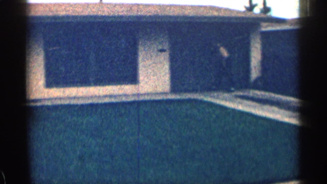 1963 Colorado home-movie frame of a low suburban house with broad dark windows, pale walls, a lawn, and heavy film-edge shadowing.