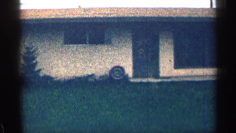 1963 Colorado frame showing a suburban house exterior with dark window openings, green grass, shrubs, and a small evergreen near the wall.