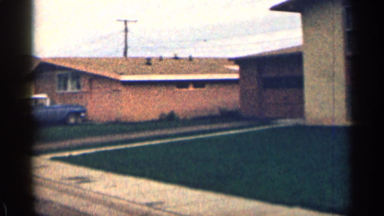 1963 Colorado home-movie frame showing a brick suburban home, green lawn, sidewalk, utility pole, and parked blue car under cloudy light.