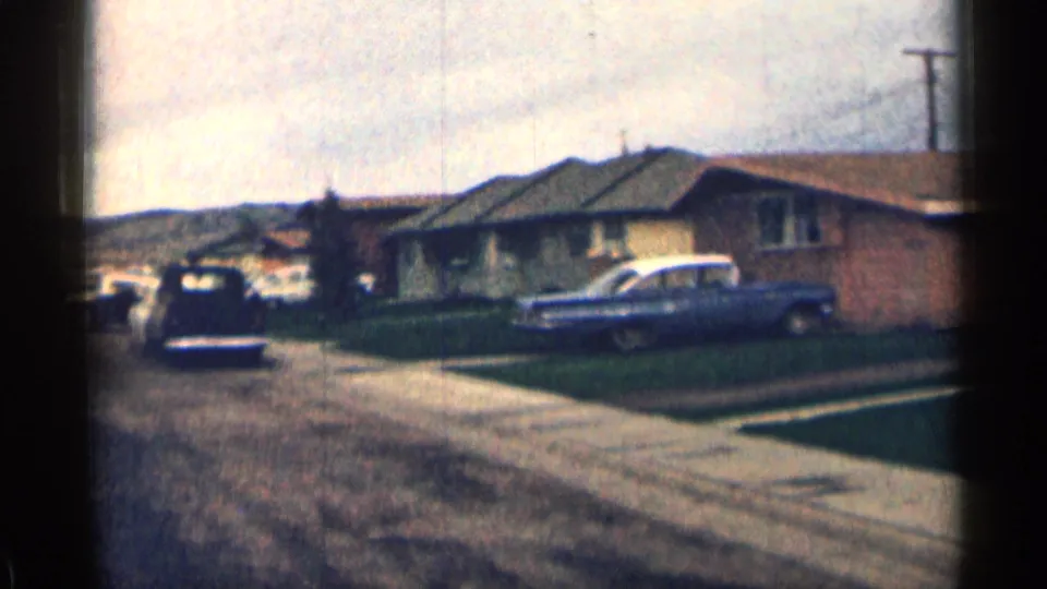 1963 Colorado suburban street scene with single-story houses, parked cars, driveways, lawns, utility poles, and low hills in the distance.