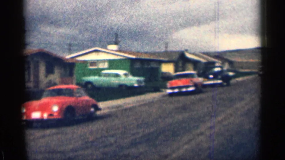 1963 Colorado home-movie frame showing a red sports car, green-sided house, pale parked car, black vehicle, lawns, and cloudy sky.