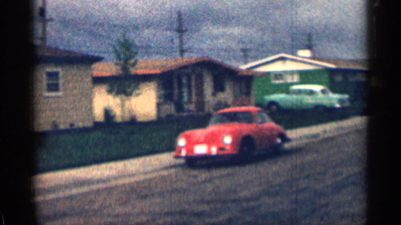 Red car passing single-story suburban homes with a pale green car parked near a driveway.