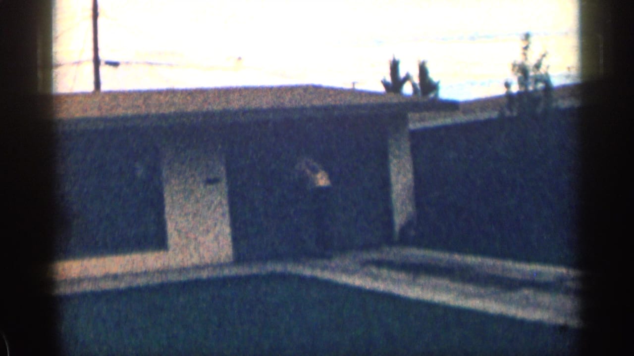 1963 Colorado home-movie frame of a dark garage or outbuilding exterior with a light door, driveway, roofline, and poles behind it.