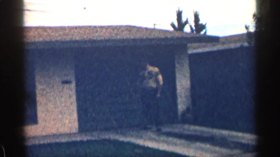 1963 Colorado home-movie frame with an adult man standing or walking near a garage exterior beside a driveway.