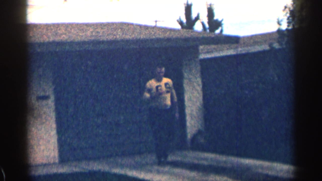 In 1963 Colorado home-movie footage, a man in a yellow number 69 jersey stands near a house exterior and dark fence.