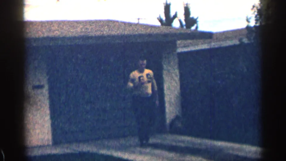In 1963 Colorado home-movie footage, a man in a yellow number 69 jersey stands near a house exterior and dark fence.