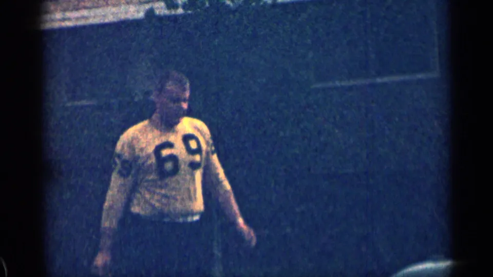 The man walks outdoors in a yellow number 69 jersey and dark pants, captured in grainy 1963 Colorado 8mm home-movie footage.