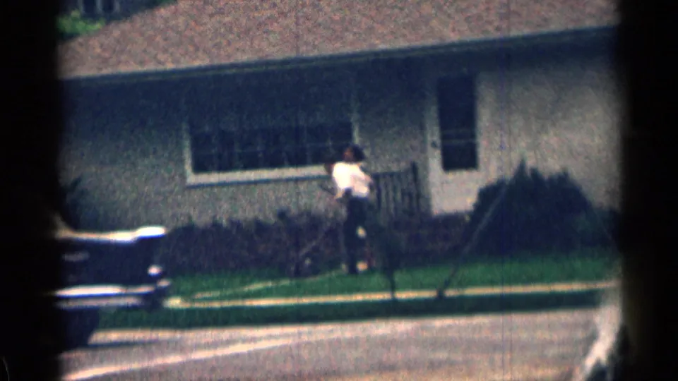 Wide 1963 Colorado home-movie view of a person near a porch, with a lawn, house facade, and parked car edge visible.