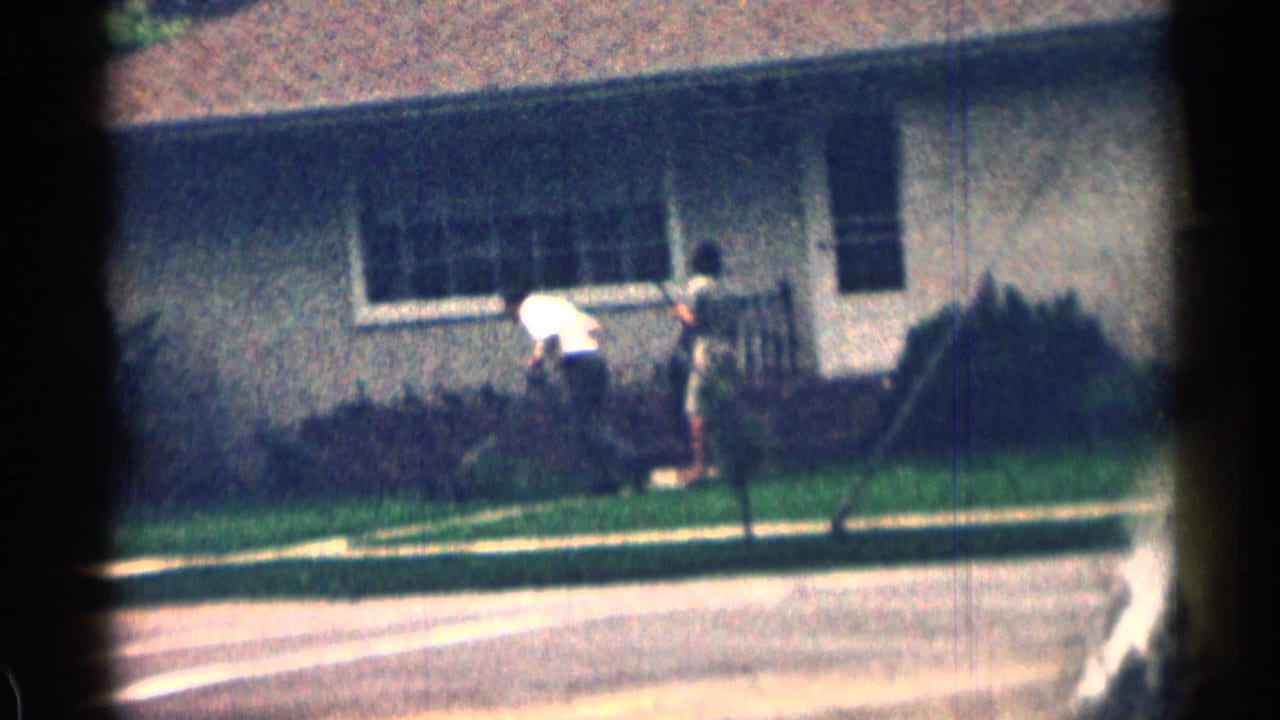 Person tending a front lawn beside a modest bungalow on a quiet residential street.