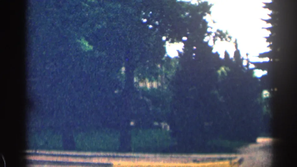 Grainy 1963 Colorado 8mm frame with trees and shrubs partly hiding a residential building beyond the street.