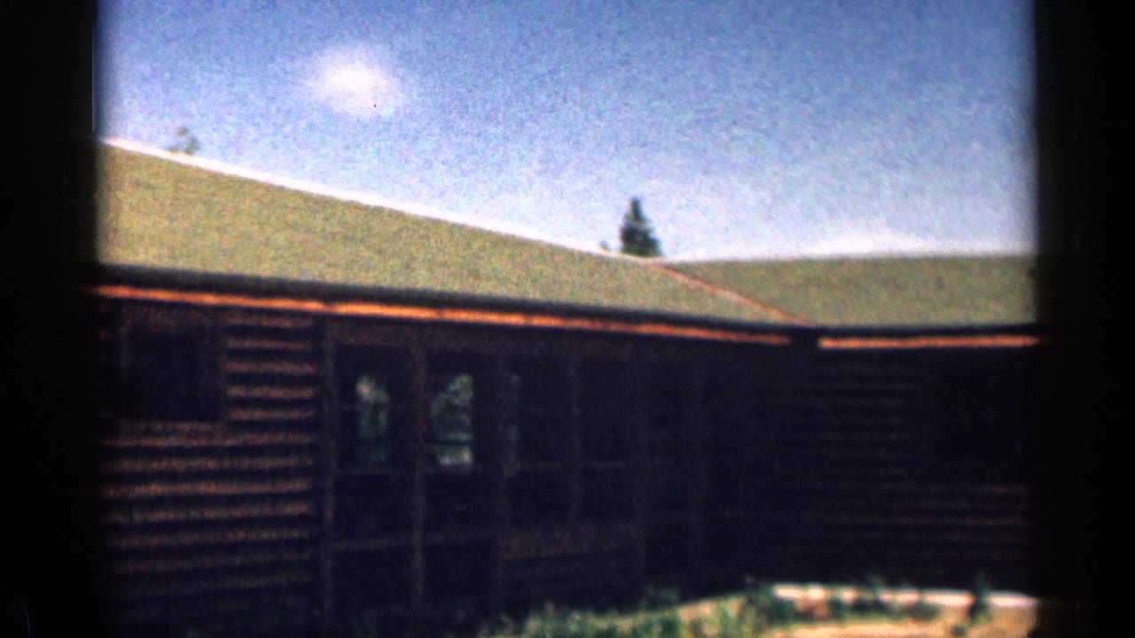 1963 Colorado home-movie frame of a rustic log cabin with a green roof, doorway, windows, and film border.