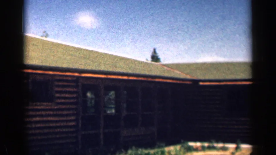 1963 Colorado home-movie frame of a rustic log cabin with a green roof, doorway, windows, and film border.