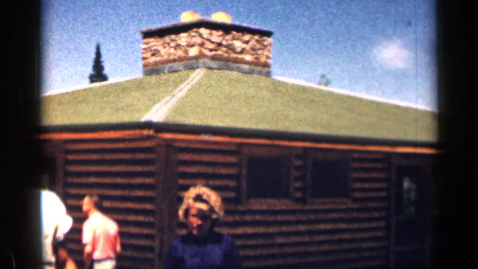 1963 Colorado frame showing visitors outside a log cabin, with a stone chimney, green roof, and bright sky.