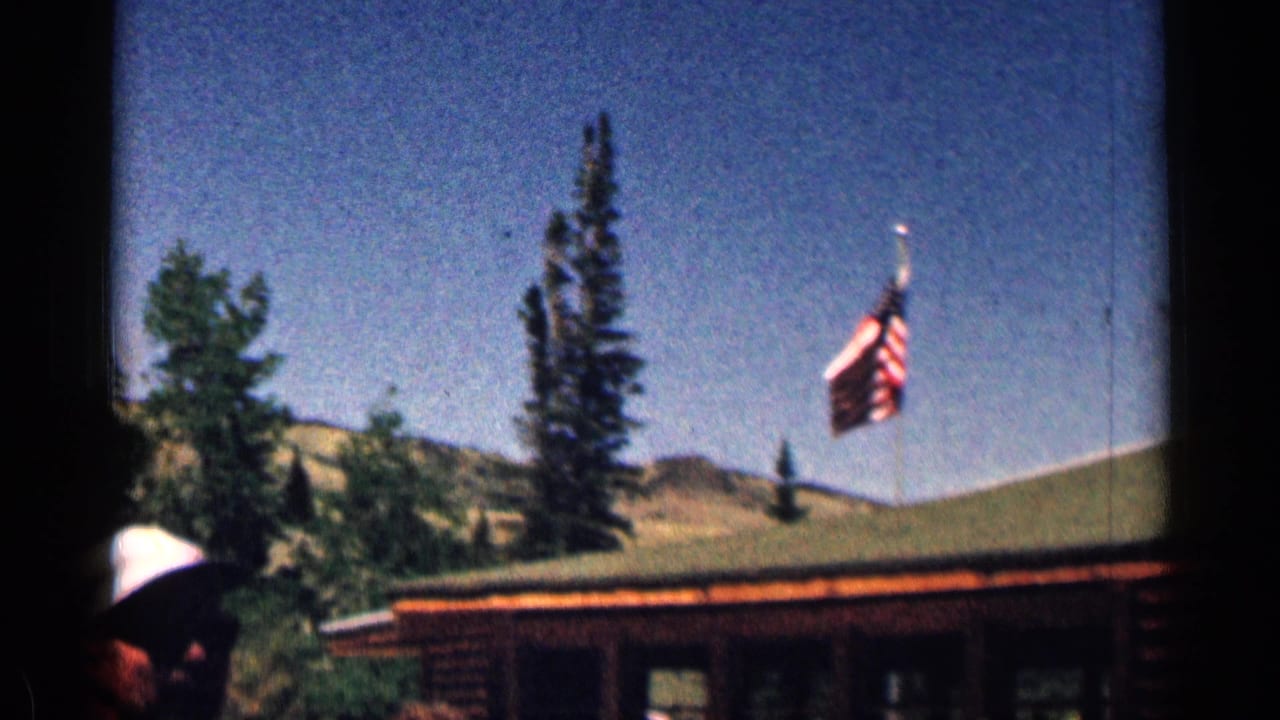 1963 Colorado 8mm frame of a rustic cabin roof with an American flag, evergreens, dry hills, and cropped visitors below.