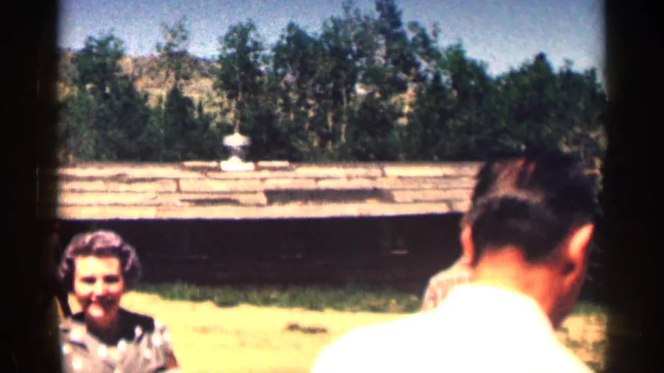 1963 Colorado home-movie frame showing adults in a mountain cabin yard, with a woman facing camera and a man in a white shirt nearby.