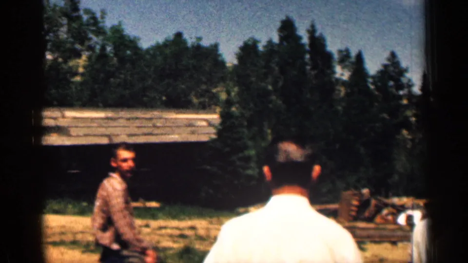 1963 Colorado cabin-yard frame with one man in a patterned shirt and another in a white shirt seen from behind among trees and logs.