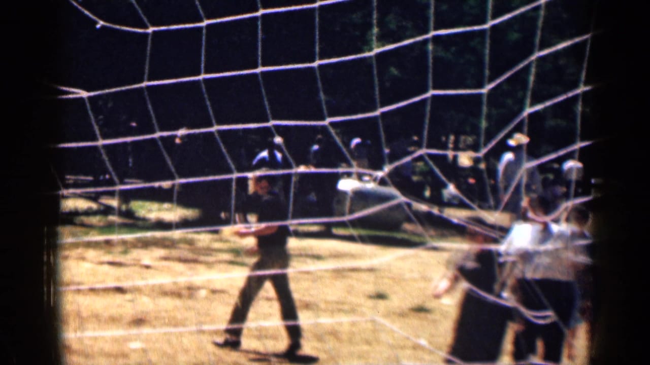 1963 Colorado 8mm frame showing a man crossing a grassy area near white netting, with several people and hills behind him.
