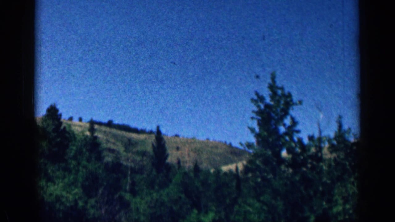Colorado mountain landscape in a 1963 8mm frame, with dark pine trees, rolling green hills, and a clear blue sky.
