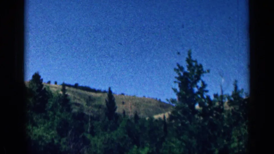 Colorado mountain landscape in a 1963 8mm frame, with dark pine trees, rolling green hills, and a clear blue sky.