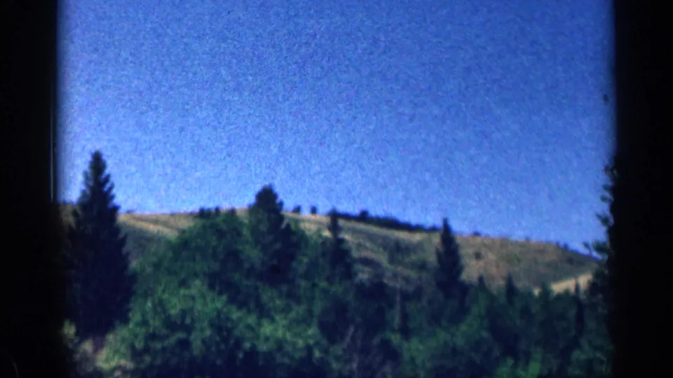 1963 Colorado 8mm landscape frame with pine trees in the foreground and rolling green hills beneath a broad blue sky.