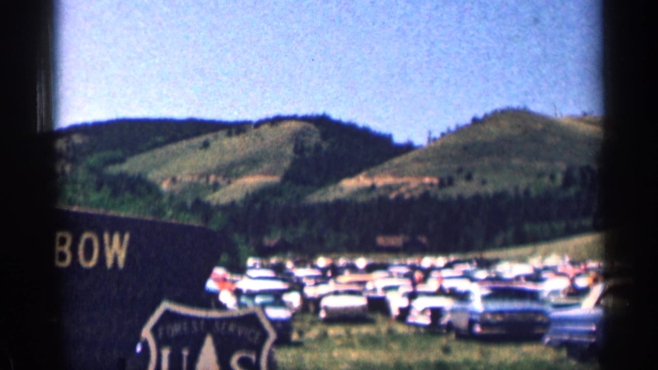 1963 Colorado home-movie frame showing rows of parked cars near forested hills and a partially visible roadside sign.