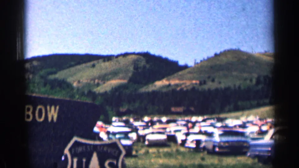 1963 Colorado home-movie frame showing rows of parked cars near forested hills and a partially visible roadside sign.
