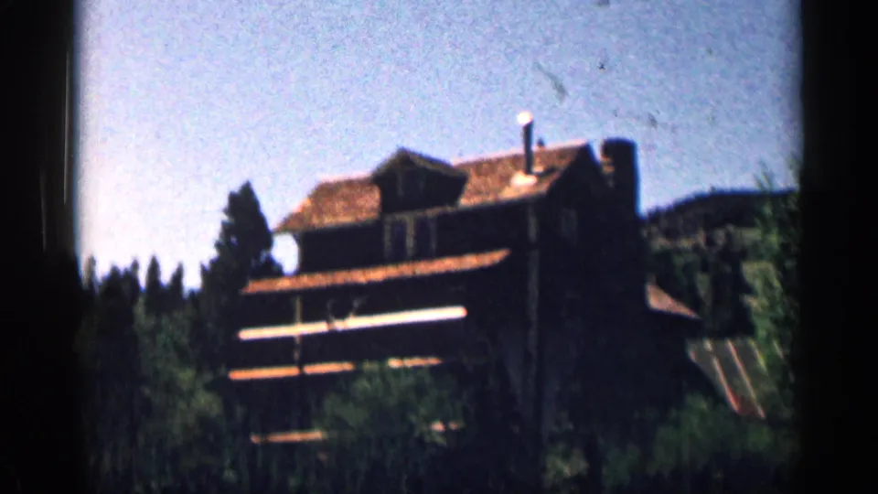 1963 Colorado 8mm frame showing a rustic mountain lodge on a wooded hillside beneath a bright blue sky.