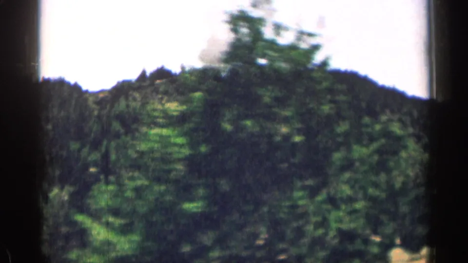1963 Colorado 8mm frame with blurred roadside evergreens in front of a pine-covered mountain ridge.