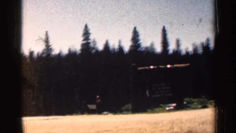A large dark entrance sign stands near a road against a line of pine trees in the Colorado home movie clip.