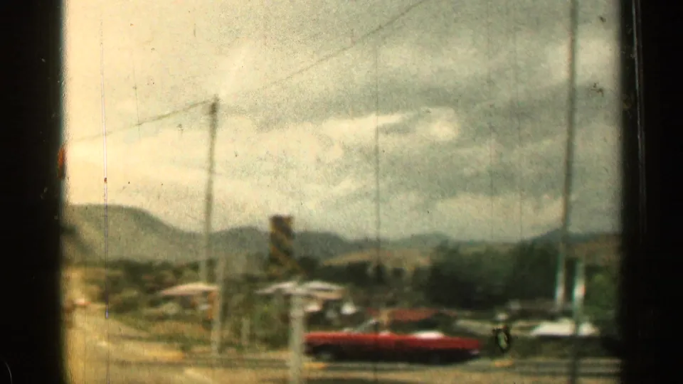 1960s road view toward a small mountain town, with utility poles, parked cars, and a red convertible visible.