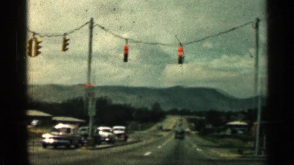 Mid-century road intersection with suspended traffic signals, cars, utility lines, and low mountains in the distance.