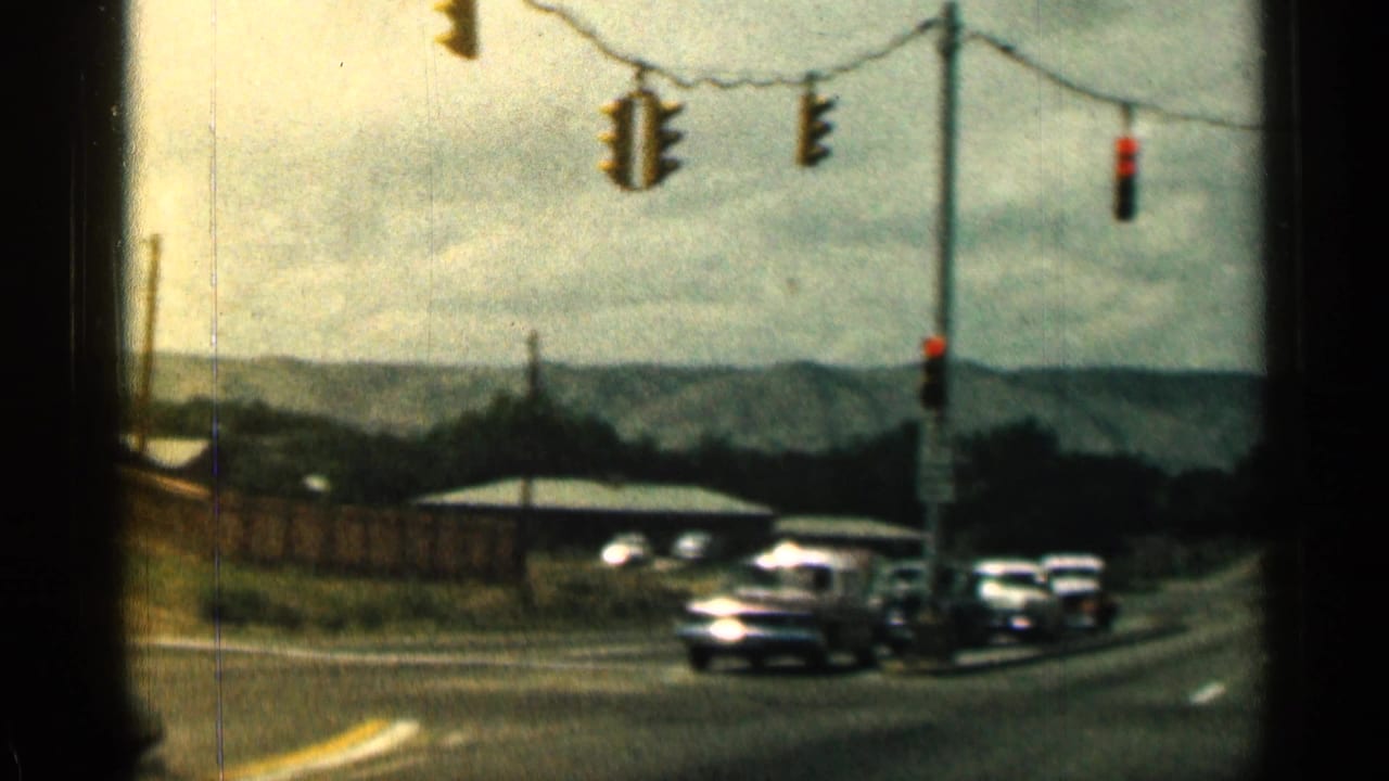 Classic cars wait at an intersection with hanging traffic lights and distant hills.