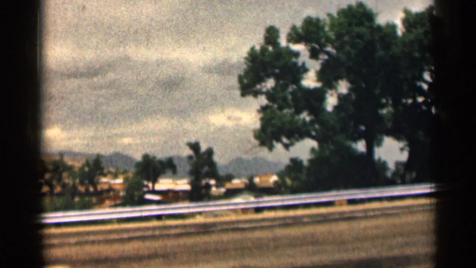 1963 Colorado road-trip frame with a guardrail, roadside trees, distant mountains, and cloudy sky seen from a moving vehicle.