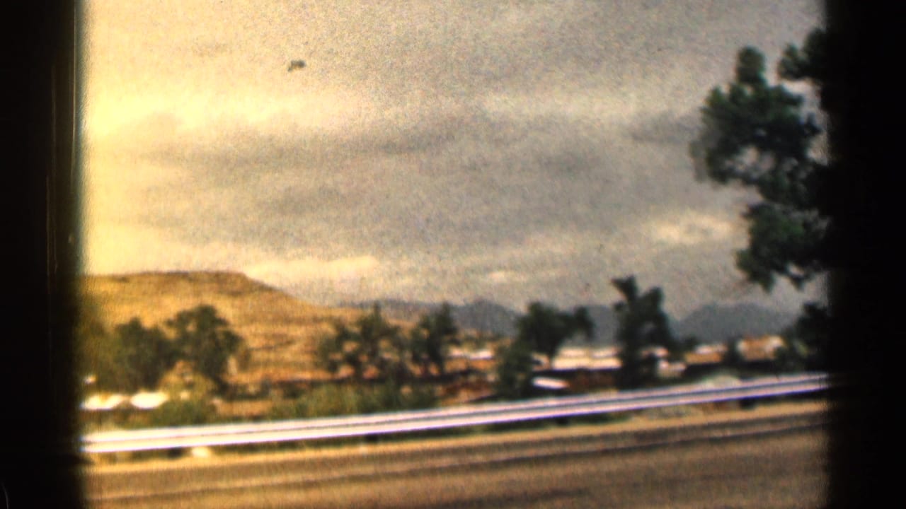 Highway and guardrail seen from a moving car with hills, trees, and cloudy sky beyond.