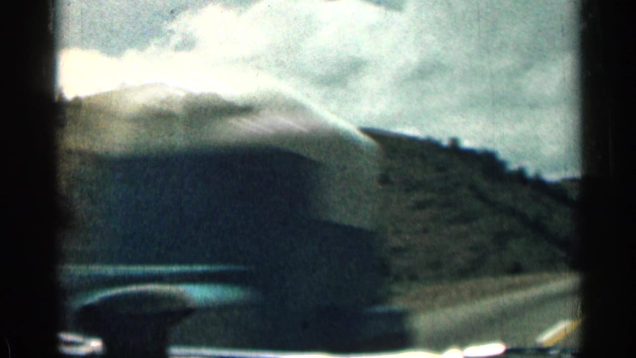 Car hood and windshield frame a hillside road scene under bright cloud cover.