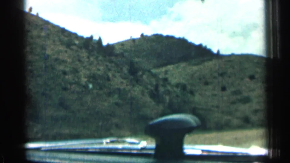 Passenger view through the windshield shows a dashboard, car hood detail, and green Colorado hills passing by on 8mm film.