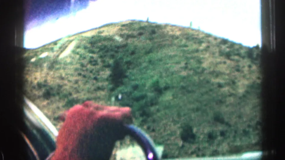 A hand rests near the steering wheel while the car moves through Colorado hills, with windshield edges and landscape visible ahead.