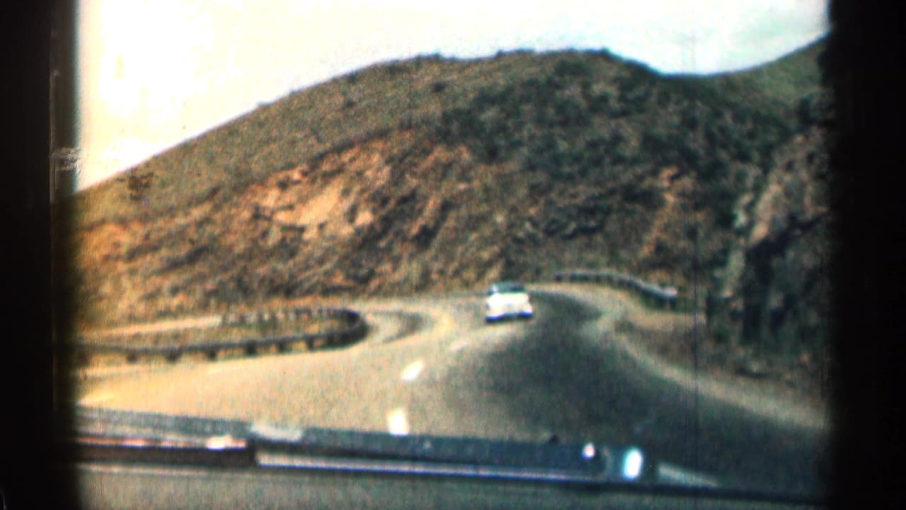 Winding mountain road seen through a car windshield with a white car ahead.