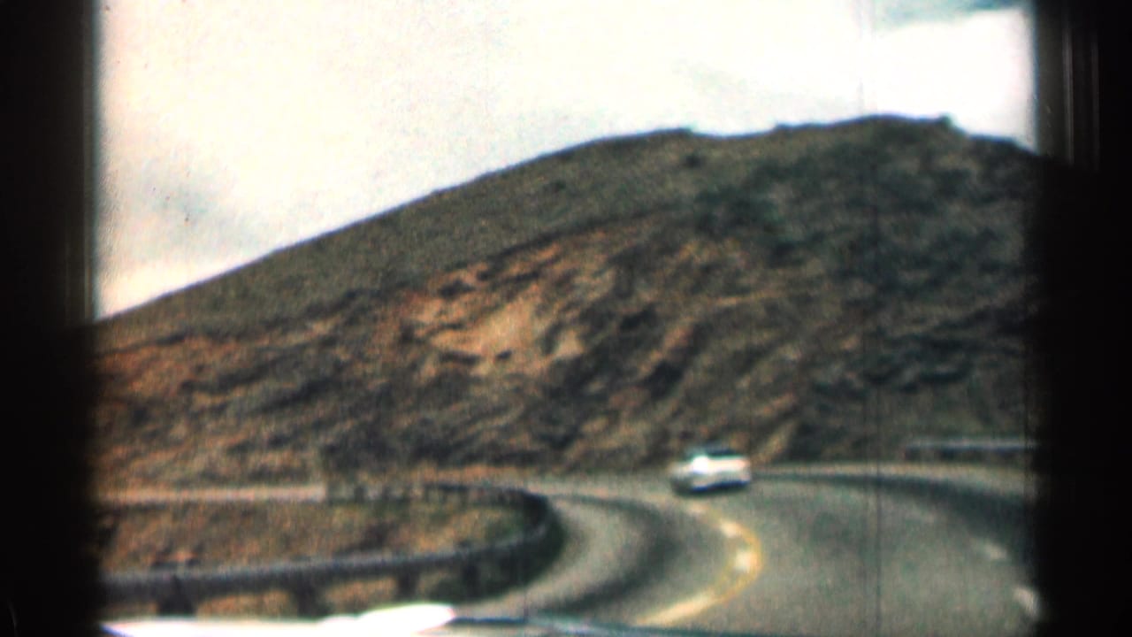 White car rounds a curve on a mountain road below rocky slopes.