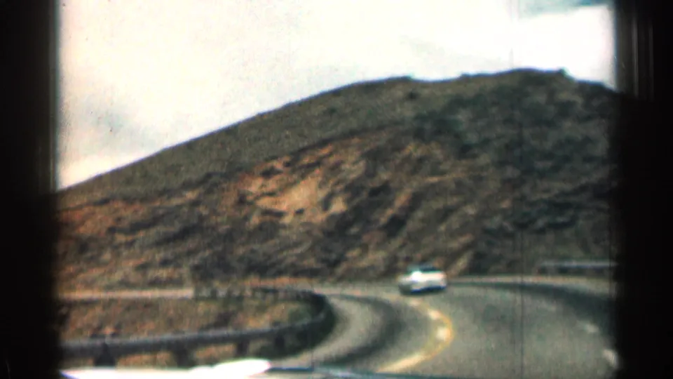 A white car travels around a bend on a Colorado mountain road, with arid slopes and guardrail visible in the 8mm frame.
