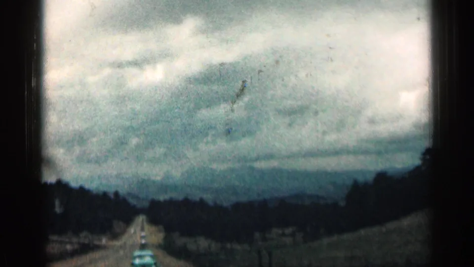 A Colorado road stretches into the distance under cloudy sky, with dark hills and roadside slopes in a soft 8mm home-movie frame.