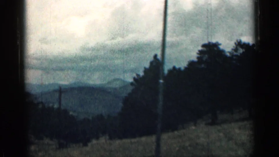 1963 Colorado roadside view from a moving car showing utility poles, dark trees, open land, and hazy mountain ridges.