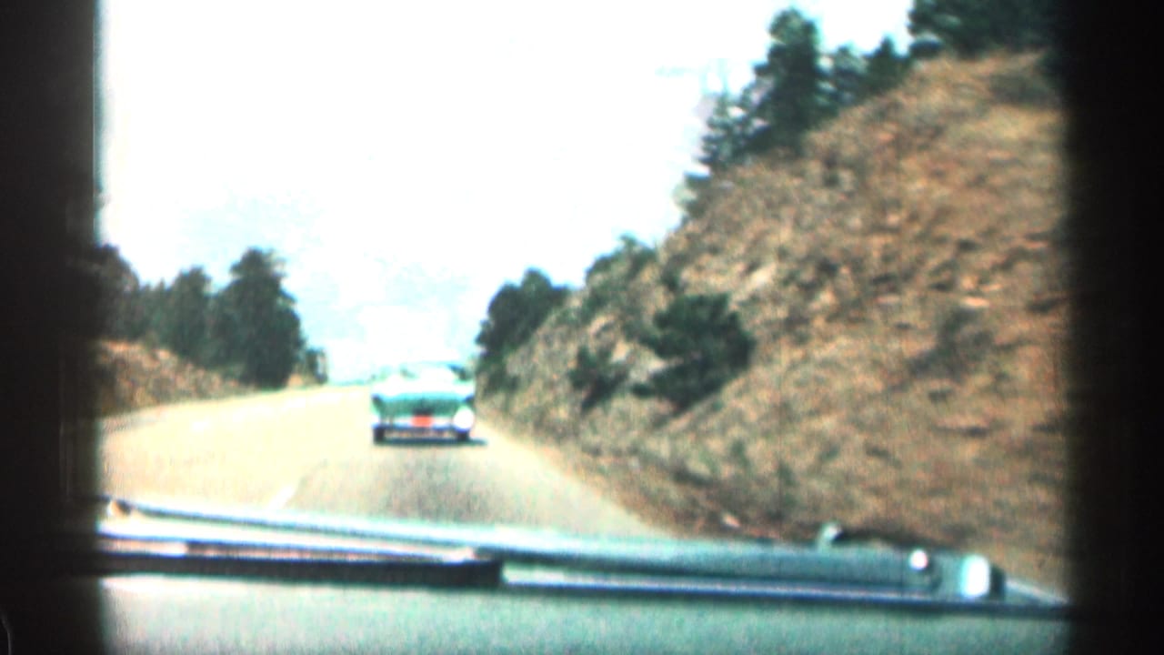 Passenger-seat view of a car following a turquoise sedan on a winding road beside rocky hills.