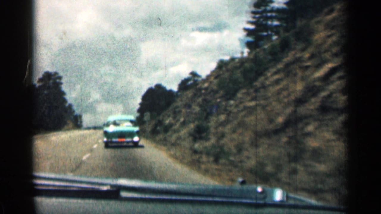 1963 Colorado road-trip footage shows a turquoise mid-century sedan ahead of the filming car on a two-lane mountain road, with rocky roadside cuts, dark tree cover, and an overcast sky.