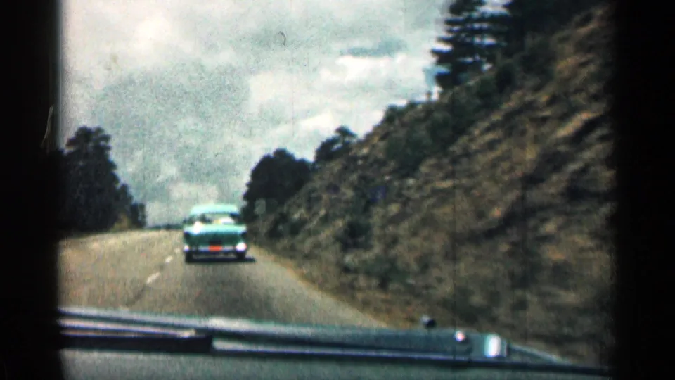 1963 Colorado road-trip footage shows a turquoise mid-century sedan ahead of the filming car on a two-lane mountain road, with rocky roadside cuts, dark tree cover, and an overcast sky.